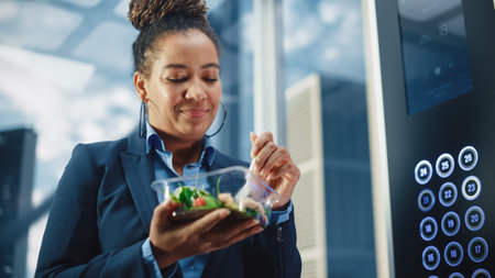 Confident Black Female Riding Glass Elevator to Office in Modern Business Center and Eating Healthy Salad with Vegetables. Happy Manager Preparing forの写真素材