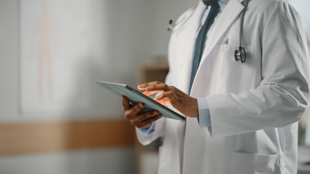 Close Up Shot of a African American Male Doctor Wearing White Coat Working on Tablet Computer at His Office. Medical Health Care Professional Workingの写真素材