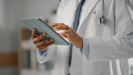 Close Up Shot of a African American Male Doctor Wearing White Coat Working on Tablet Computer at His Office. Medical Health Care Professional Workingの写真素材