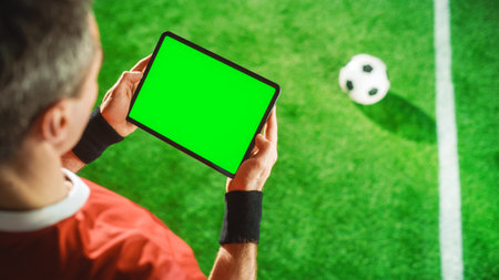Professional Soccer Player Holding Digital Tablet Computer with Green Screen Chroma Key Template. Elevated Top Down Above Shot of Football Star Browsing Content While Standing on Game Fieldの写真素材