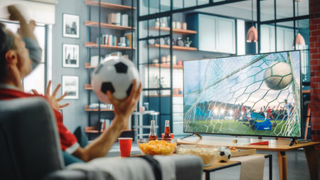 At Home Soccer Fans Sitting on a Couch Watch Football Game on TV, Cheer for Favourtite Sports Team to Win Championship. Screen Shows Professional Football Club Play. Over the Shoulderの写真素材