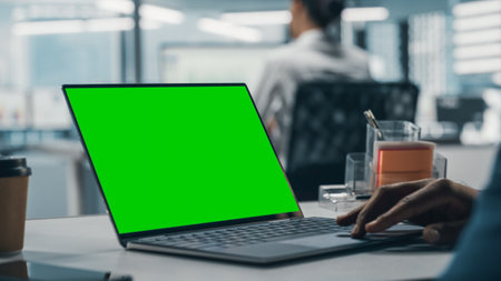 Successful Black Businessman Sitting at Desk Working on Green Screen Laptop in Office. African American Businessperson using Touch Pad on Chroma Key Computer. Shot with Focus on Handの写真素材