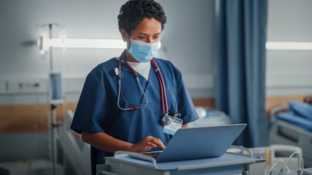 Hospital Ward: Professional Black Latin Female Head Nurse or Doctor Wearing Face Mask Uses Medical Computer. In the Background Patients in Bedsの写真素材