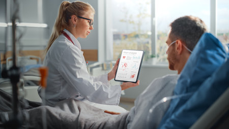 Hospital Ward: Friendly Female Doctor Talks to Sick Male Patient Resting in Bed, Uses Tablet Computer, Shows and Explains Heart Scan Resultsの写真素材