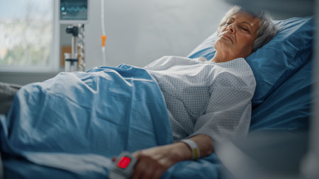 Hospital Ward: Senior Woman Resting in a bed with Finger Heart Rate Monitor / Pulse oximeter showing Pulse. Her Fragile Hands Resting on a Blanketの写真素材