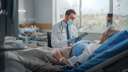 Hospital Ward: Friendly Doctor Wearing Face Mask Talks to Beautiful Senior Female Patient Resting in Bed, Explains Test Results, Gives Recovery Adviceの写真素材