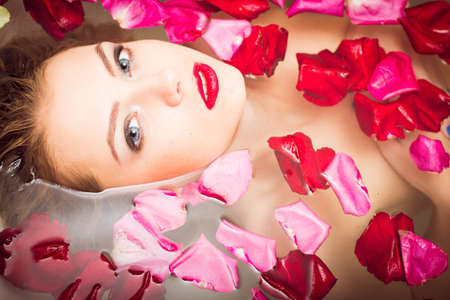 luxury spa: elegant blue eyes lady pinup girl with silk skin having fun happy relaxing laying in water bath & looking at camera on colorful rose flower petals copy space background close up portraitの写真素材