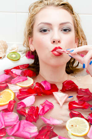 Icecream in spa: closeup portrait of beautiful young tempting lady eating ice cream in bath with rose petals and fruit slices. luxury happy girl happy smiling & looking at cameraの写真素材