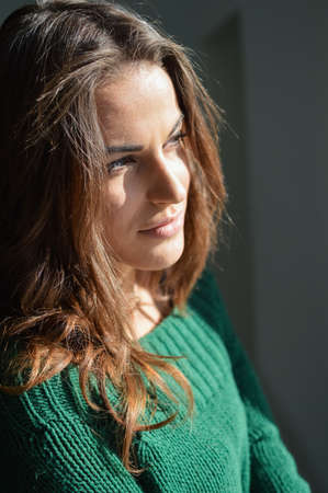 closeup portrait of beautiful young woman in woolen sweater enjoying sunshine and looking out the windowの写真素材