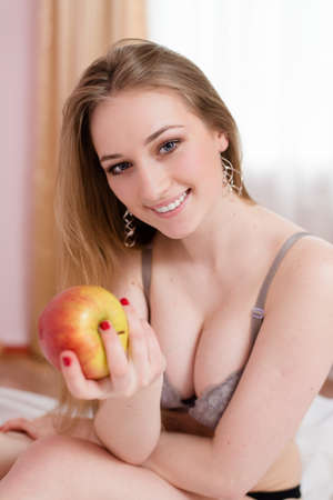 portrait of sexy blonde girl having fun happy smiling holding big red apple sitting on white bed in lingerie and looking at camera on light window copy space backgroundの写真素材