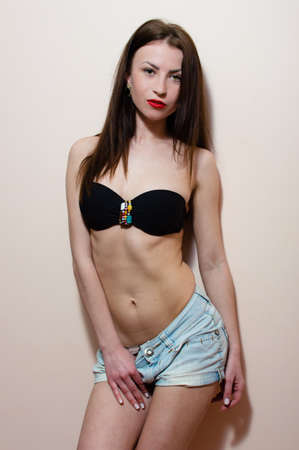 portrait of brunette fashion sexy young lady with red lips in jeans shorts showing perfect fit stomach & looking at camera on white or light wall copy space backgroundの写真素材