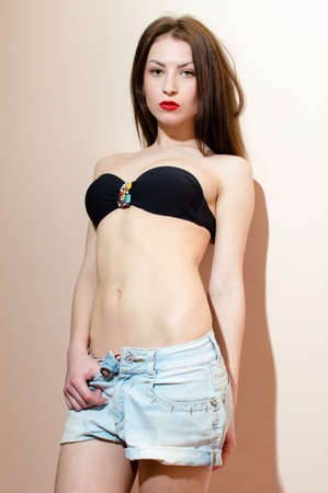 brunette fashion sexy girl with red lips in jeans shorts showing perfect fit stomach & looking at camera, portrait on white or light wall copy space backgroundの写真素材