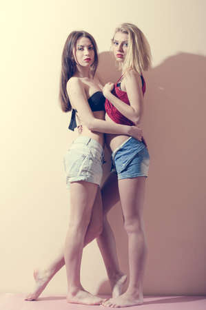 2 fashion sexy romantic beautiful girls in jeans shorts having fun & good time hugging each other, portrait on light copy space backgroundの写真素材