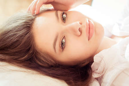 happy looking at camera young beautiful charming woman in white shirt lying on the bed, closeup portraitの写真素材