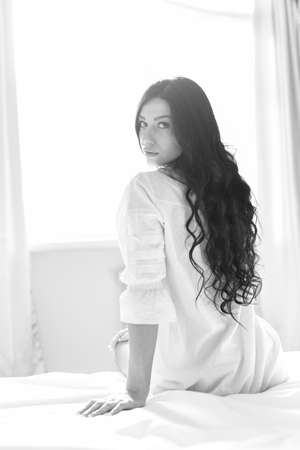 picture of brunette pretty girl or woman looking at camera and sitting on white bed on a sunny window copy space backgroundの写真素材