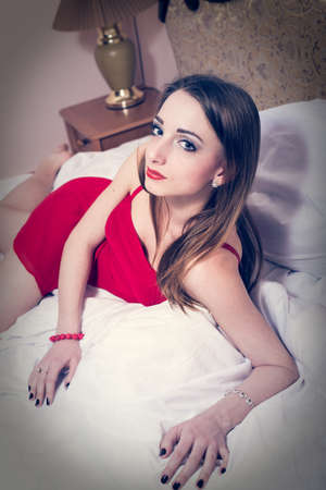 portrait of elegant sensual beautiful young lady having fun in red dress lying on the white bed on copy space bedroom backgroundの写真素材