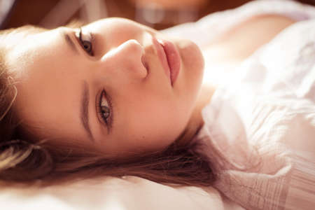 closeup portrait of happy looking at camera young beautiful charming woman in white shirt lying on the bedの写真素材