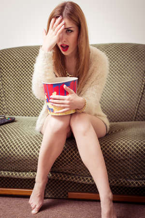portrait of young blond pretty lady with popcorn sitting on sofa watching tv and hiding face with palm up on light copy space backgroundの写真素材