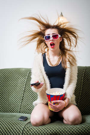 great movie moments: portrait of funny blond pretty girl having fun watching tv movie in 3d glasses with popcorn holding remote control & mouth wide open on light copy space backgroundの写真素材