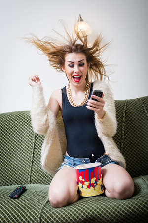 great news: portrait of funny young blond pretty woman having fun watching tv movie with popcorn and holding mobile phone, excited happy screaming on light copy space backgroundの写真素材