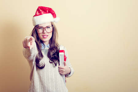 portrait of pointing finger & holding present box beautiful young lady in Santa Claus hat, glasses happy smiling and looking at cameraの写真素材