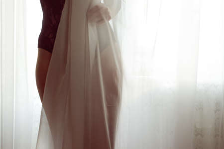 closeup on silhouette of hiding behind tulle curtain pretty lady with perfect fit body in black combies dress standing on light window copy space backgroundの写真素材