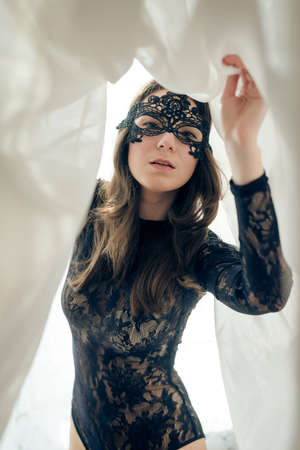portrait of young beautiful lady with perfect fit body in black combies dress & mask standing on light curtain copy space backgroundの写真素材