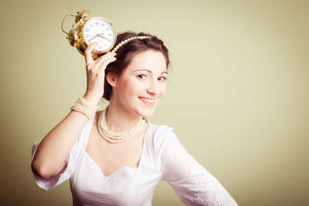 portrait of elegant beautiful young lady in white dress having fun holding clock up on head & happy smile on copy space backgroundの写真素材