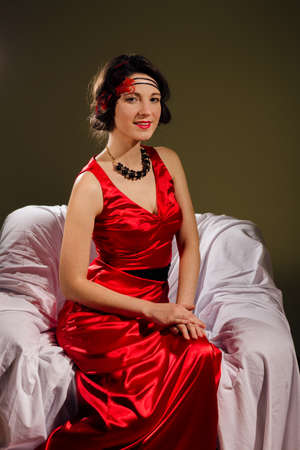 portrait of aristocracy retro styled beautiful young lady in red dress relaxing sitting in chair happy smile and looking at camera on copy space backgroundの写真素材