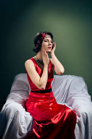 portrait of emotional retro styled beautiful young lady in red dress relaxing sitting in chair on copy space backgroundの写真素材