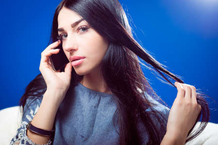 closeup portrait of pretty young lady talking on mobile phone and sensually looking at camera while touching luxury hair on blue copy space backgroundの写真素材