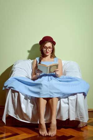 Girl in glasses and red hat sitting on the couch reading a bookの写真素材