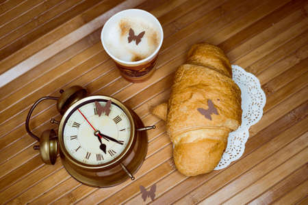 Image of alarm clock, cup of hot drink and croissant