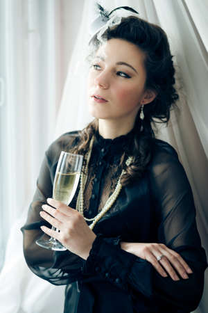 Portrait of beautiful woman in black dress and glass of champagne and looking out the windowの写真素材