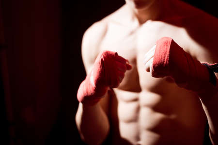 Young man training box punches with bandages closeup on strong muscles of torso over dark studio...