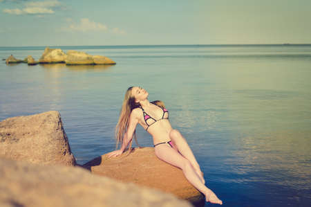 Picture of sexi young blond female in swimwear with long beautiful hair sunbathing on rock over sea backgroundの写真素材