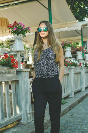 Portrait of carefree young pretty lady with long hair in sunglasses and hands in pockets walking downdown along cobbled path with potted flowering plants on background.の写真素材