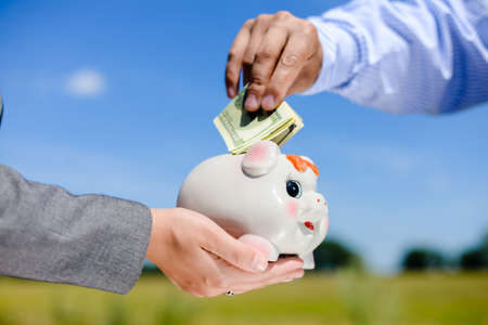 Closeup of hands holding a piggy bank with a dollar bills stuck in the top slot. Side view in horizontal format over a blue sky sunny outdoors background.の写真素材