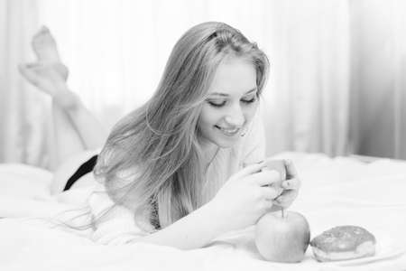 Beautiful sexi blonde young lady lying in bed and happy smiling looking at plate with donut and apple on light window background. Black and white closeup portraitの写真素材
