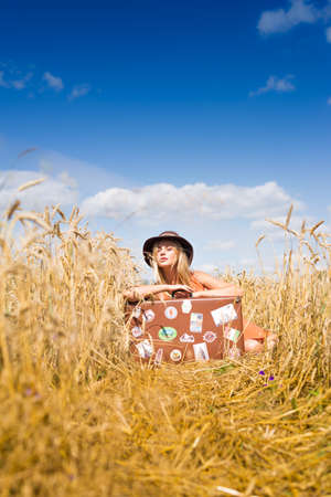 Young beautiful romantic woman in hat sitting in field of wheat with retro suitcase over blue sky on summer day outdoors backgroundの写真素材
