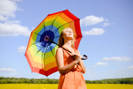 Picture of young beautiful lady in orange sundress under colorful umbrella standing over rural field with closed eyes, on sunny day outdoors backgroundの写真素材