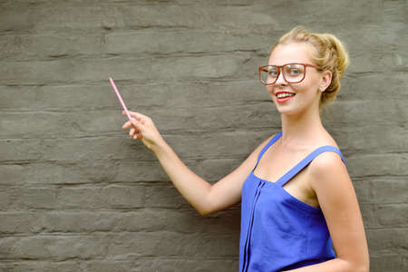 Copyspace picture of young smiling pretty lady in glasses pointing to the brickwall with pen....