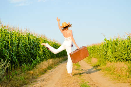 Happy jumping girl wearing hat with leather suitcase on road in field. Backview of happy woman in white costume.の写真素材