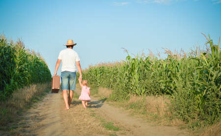 Man and babygirl walking away on road between corn field over blue sky outdoors background. Backview of father carrying suitcase with daughter.の写真素材