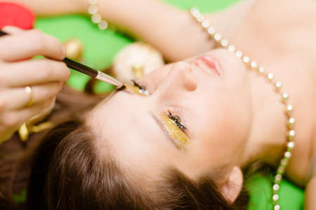 Portrait closeup of applying makeup on face of lying brunette girl. Golden eye makeup for woman lying on blurred green background.の写真素材