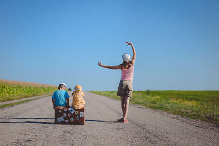 Picture of young woman standing inn ballet pose and two children sitting on old suitcase on country road. Backview of family traveling on blue sky sunny outdoors background.の写真素材