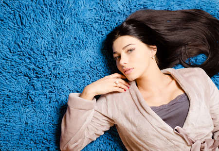 Picture of beautiful woman in beige robe lying on blue fluffy carpet. Young girl looking at camera on bright comfort indoor background.の写真素材