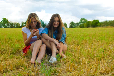 Picture of two pretty young women with long hair sitting on summer meadow. One girl smiling while another typing and reading on mobile phone on outdoor background.の写真素材