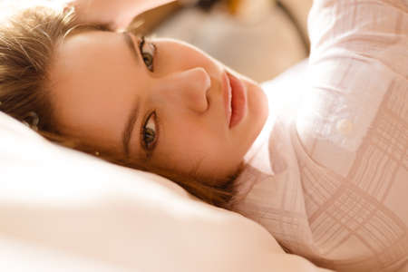Portrait of sexy beautiful charming girl lying on white pillow in morning sunlight. Closeup of young lady looking in camera on blurred sunny indoor background.の写真素材