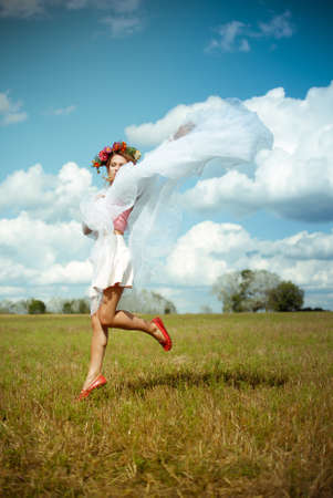 Picture of amazing beautiful girl in flower wreath with white chiffon shawl dancing on summer meadow. Charming woman in red shoes on sunny countryside background.の写真素材